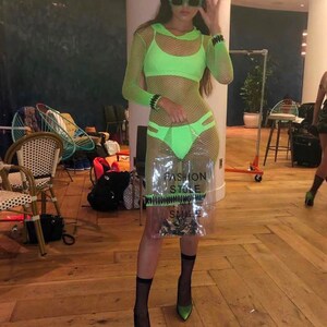 May include: A woman wearing a neon green mesh dress with a matching bra top and black fishnet socks. She is holding a clear plastic bag with the text "Fashion Stole My Smile" printed on it. She is wearing green sunglasses and black heels.