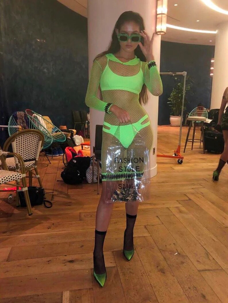 May include: A woman wearing a neon green mesh dress with a matching bra top and black fishnet socks. She is holding a clear plastic bag with the text "Fashion Stole My Smile" printed on it. She is wearing green sunglasses and black heels.