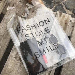 May include: Clear plastic tote bag with a black handle and the text "FASHION STOLE MY SMILE" printed on the front. The bag contains sunglasses, a black wallet, and a red lipstick.