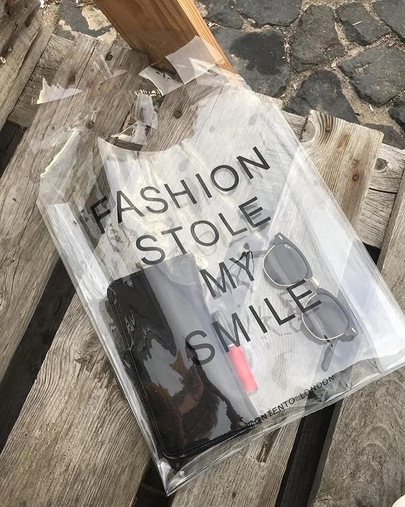 May include: Clear plastic tote bag with a black handle and the text "FASHION STOLE MY SMILE" printed on the front. The bag contains sunglasses, a black wallet, and a red lipstick.