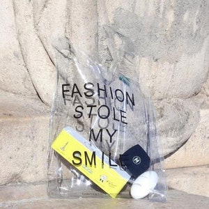 May include: A clear plastic tote bag with the text "FASHION STOLE MY SMILE" printed on it. The bag contains a yellow tube of hand cream, a black and white patterned soap bar, and a black hat with a white logo.
