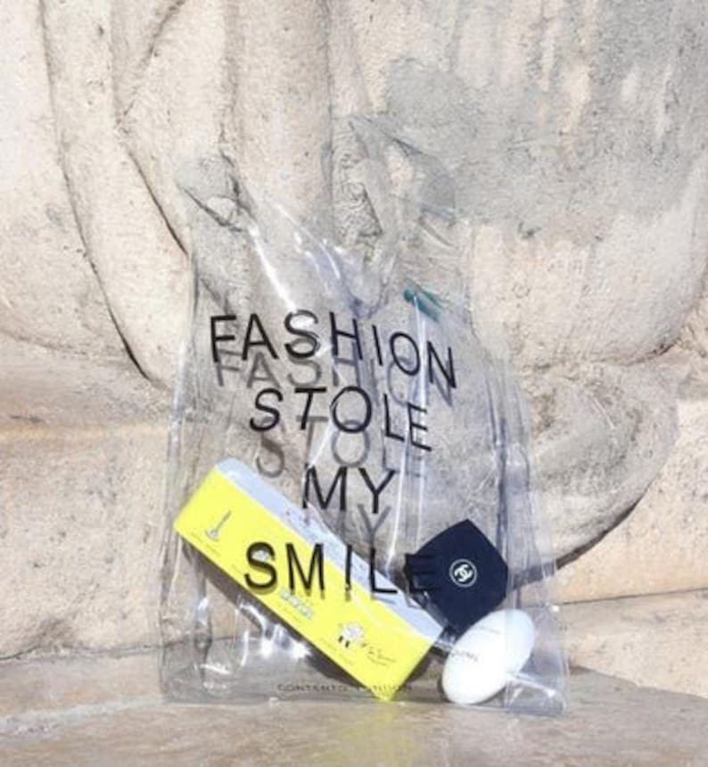 May include: A clear plastic tote bag with the text "FASHION STOLE MY SMILE" printed on it. The bag contains a yellow tube of hand cream, a black and white patterned soap bar, and a black hat with a white logo.