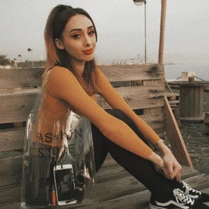 May include: A young woman wearing a brown long-sleeved shirt and black pants sits on a wooden bench. She is holding a clear plastic bag with the text "Fashion Store" printed on it. The bag contains a black phone and a silver object.