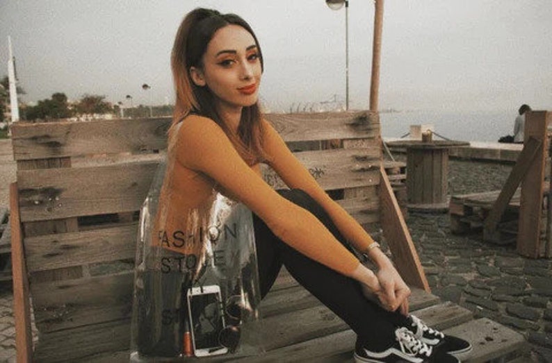 May include: A young woman wearing a brown long-sleeved shirt and black pants sits on a wooden bench. She is holding a clear plastic bag with the text "Fashion Store" printed on it. The bag contains a black phone and a silver object.