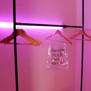 May include: Pink room with a black clothing rack and pink hangers. A clear plastic bag hangs from the rack with the text "FASHION STOLE MY SMILE".