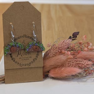 May include: A pair of dangle earrings featuring a semi-circular brown house design with a central circular door and green and purple foliage accents. The earrings are attached to a brown cardboard display card with the text A Moon Holiday Decor Kate Kish.