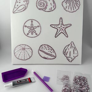 May include: A white canvas featuring eight sea-themed designs outlined in small pink rhinestones. Below the canvas are a purple plastic tray, a tube of B-7000 adhesive, a purple applicator pen, a small square of wax, and three bags of pink rhinestones.