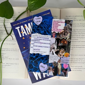 Puede incluir: Una composición que incluye un libro, dos marcapáginas decorativos y una planta. El libro tiene una cubierta azul con el título "TAM" y las palabras "IN A LIFE OF STORMS, THE SUN" en blanco. Los marcapáginas tienen texto e imágenes.