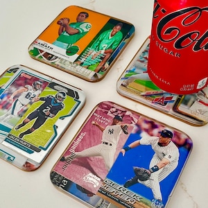 May include: Six different sports coasters featuring various athletes. The coasters are made of a clear material with a printed image on top. The coasters are arranged in a circle around a can of cola.
