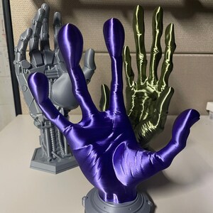 May include: Three 3D printed hands, one gray, one green, and one purple. The hands are posed in a variety of positions, with the purple hand raised in the air.