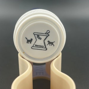 K&ouml;nnte beinhalten: Wei&szlig;er Plastikflaschenverschluss mit einem schwarz-wei&szlig;en Logo. Das Logo zeigt einen M&ouml;rser und einen St&ouml;&szlig;el mit dem Text "Rx Veterinary" und Silhouetten einer Katze und eines Hundes.