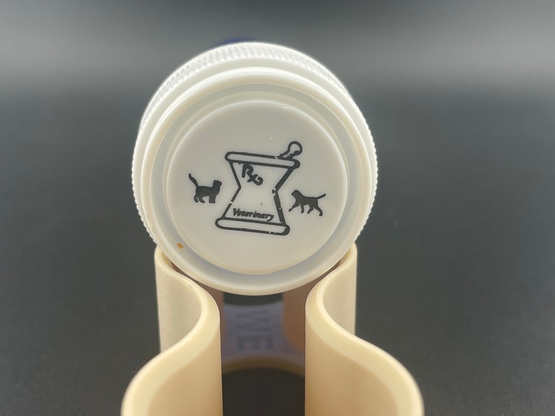 K&ouml;nnte beinhalten: Wei&szlig;er Plastikflaschenverschluss mit einem schwarz-wei&szlig;en Logo. Das Logo zeigt einen M&ouml;rser und einen St&ouml;&szlig;el mit dem Text "Rx Veterinary" und Silhouetten einer Katze und eines Hundes.