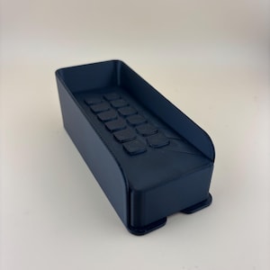 May include: A navy blue rectangular tray with a series of square indentations on the inside. The tray has a curved front and sits on two small feet. The object is made of a solid, dark material.