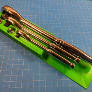 May include: A set of three chrome ratchets of varying sizes, neatly arranged in a bright green holder. The tools are positioned diagonally, showcasing their polished metal finish. The holder is on a blue grid surface.