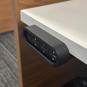 May include: A close-up of a black control panel with buttons labeled with numbers and arrows, mounted on the edge of a white desk. The desk is next to a wooden cabinet. The control panel is for adjusting the height of the desk.