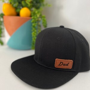 Op de afbeelding: Een zwarte en bruine baseballpet met een leren patch waarop "Dad" staat.