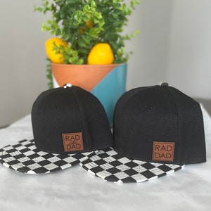 Op de afbeelding: Twee zwarte baseballpetten met een geruit patroon op de klep. De petten hebben een bruin leren patch met de tekst "RAD like DAD" erop.
