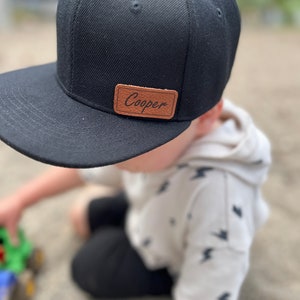 May include: A black and brown baseball cap with the name "Cooper" on a brown leather patch. The cap is worn by a child wearing a white shirt with a lightning bolt pattern.