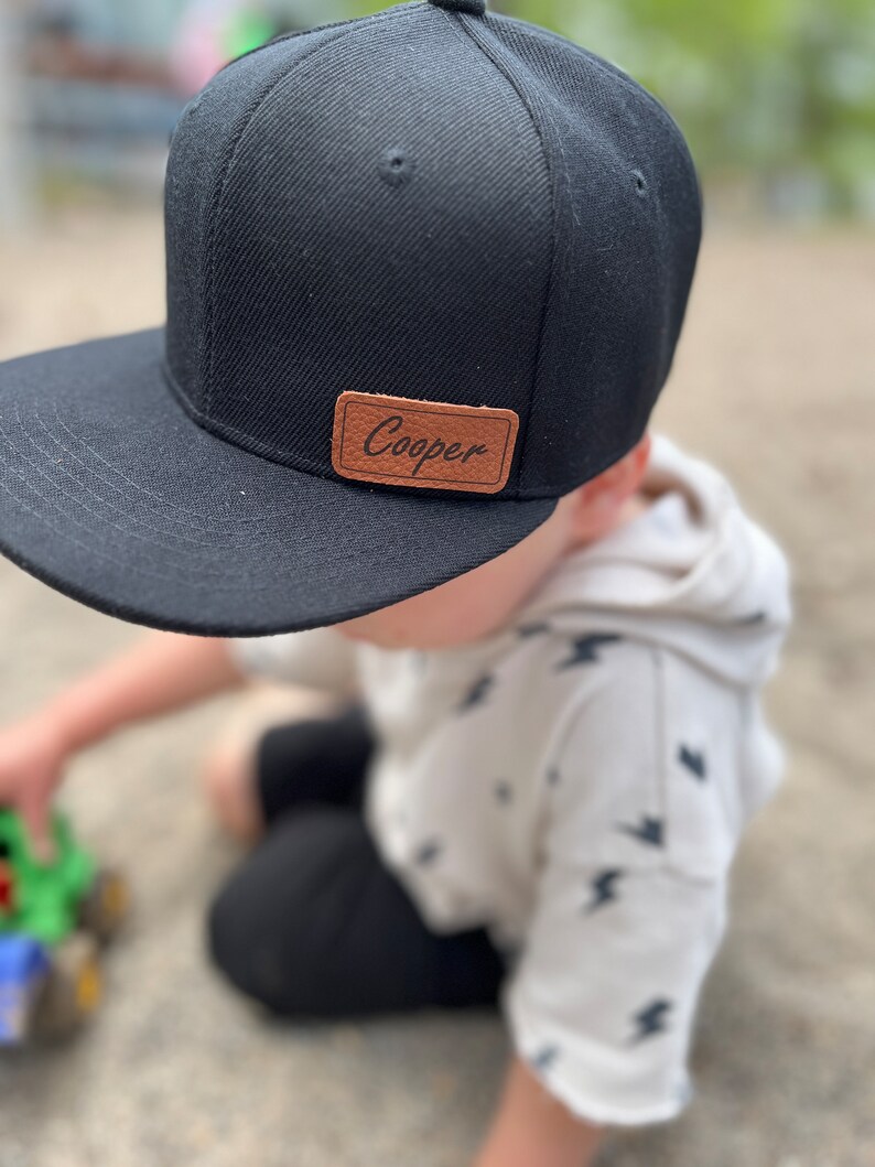 May include: A black and brown baseball cap with the name "Cooper" on a brown leather patch. The cap is worn by a child wearing a white shirt with a lightning bolt pattern.