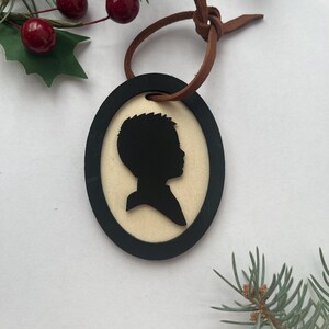 May include: Oval-shaped wooden ornament with a black silhouette of a person's profile. The silhouette is set against a light-colored background and framed by a black border. A brown leather cord is attached for hanging. Decorative sprigs and berries are in the background.