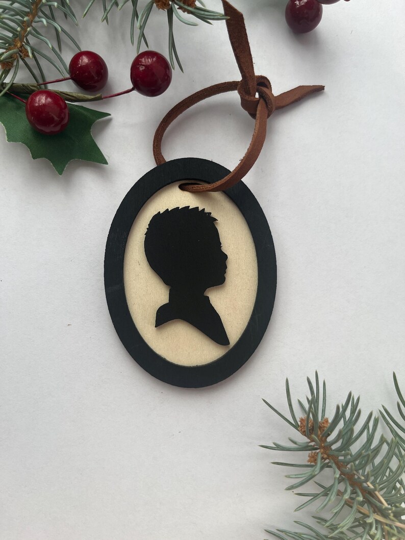 May include: Oval-shaped wooden ornament with a black silhouette of a person's profile. The silhouette is set against a light-colored background and framed by a black border. A brown leather cord is attached for hanging. Decorative sprigs and berries are in the background.