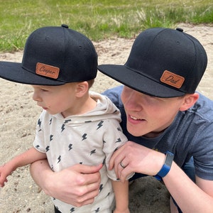 May include: A father and son wearing black baseball caps with brown leather patches. The father's patch says "Dad" and the son's patch says "Cooper".