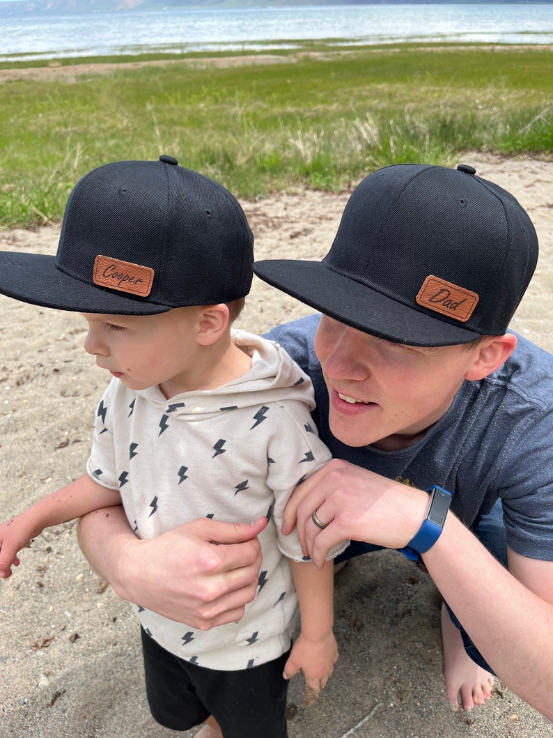 May include: A father and son wearing black baseball caps with brown leather patches. The father's patch says "Dad" and the son's patch says "Cooper".
