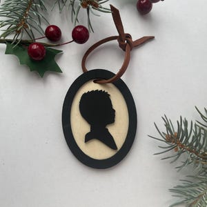 May include: Oval-shaped Christmas ornament featuring a black silhouette of a person's profile. The silhouette is set against a light-colored background, framed by a black border. A brown leather cord is attached for hanging. Festive holiday decor.
