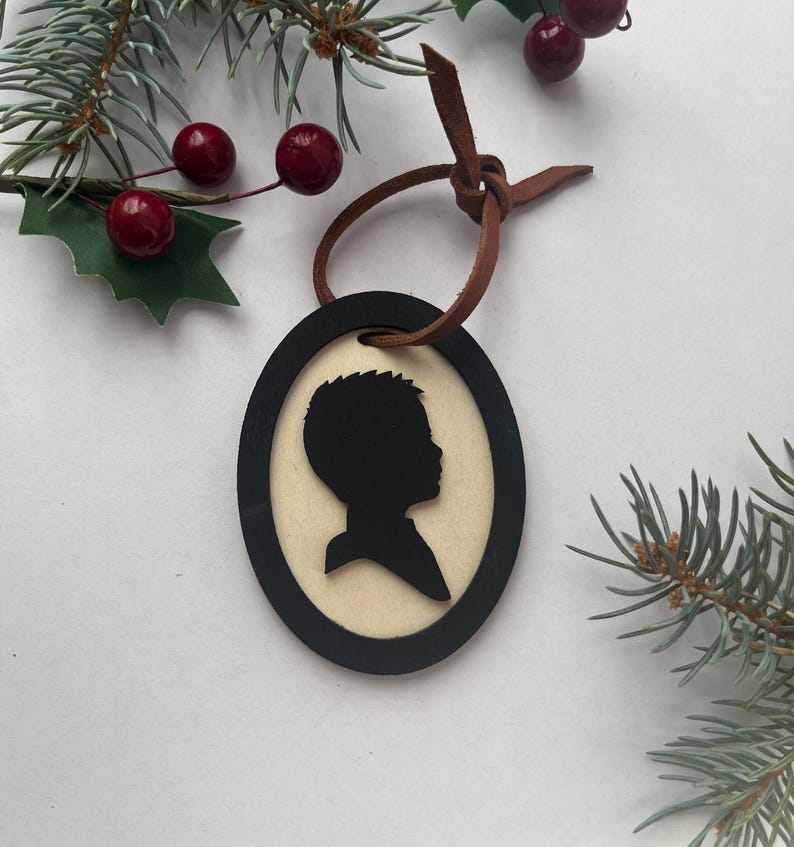 May include: Oval-shaped Christmas ornament featuring a black silhouette of a person's profile. The silhouette is set against a light-colored background, framed by a black border. A brown leather cord is attached for hanging. Festive holiday decor.