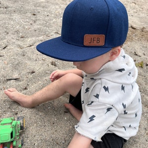 May include: A young child wearing a navy blue baseball cap with a brown leather patch that says "JFB" on it. The child is sitting on a sandy surface and wearing a white shirt with a black lightning bolt pattern.