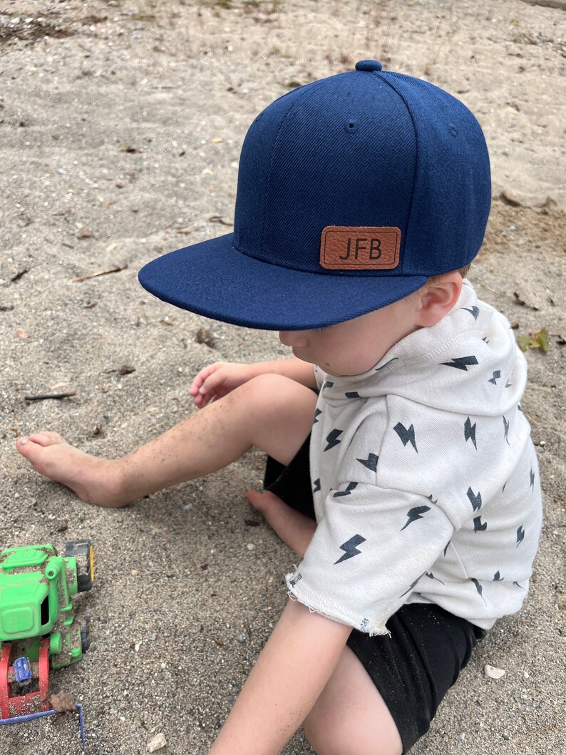 May include: A young child wearing a navy blue baseball cap with a brown leather patch that says "JFB" on it. The child is sitting on a sandy surface and wearing a white shirt with a black lightning bolt pattern.