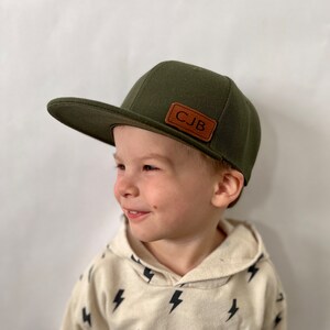 May include: A young child wearing a green baseball cap with a brown leather patch that says "CJB". The child is wearing a white shirt with a black lightning bolt pattern.