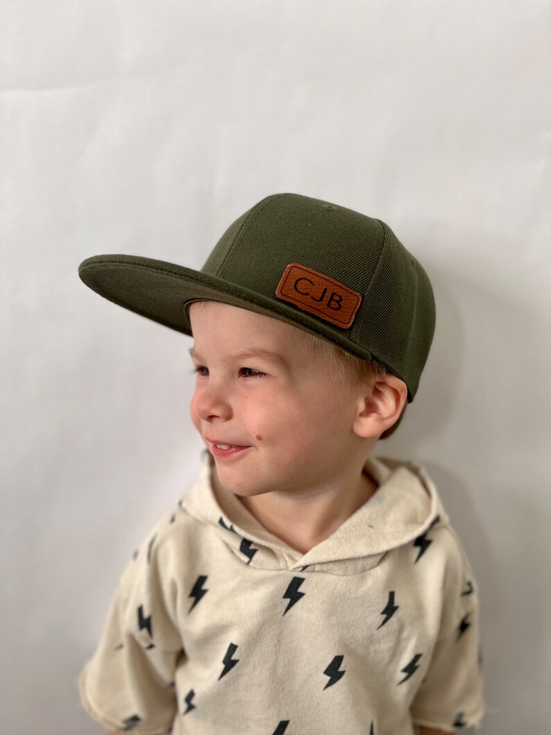 May include: A young child wearing a green baseball cap with a brown leather patch that says "CJB". The child is wearing a white shirt with a black lightning bolt pattern.