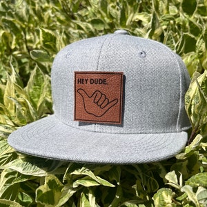 Pode incluir: Um boné de beisebol cinza e branco com um remendo de couro marrom que diz "HEY DUDE." O remendo apresenta um contorno preto de uma mão fazendo o sinal shaka.