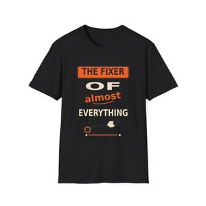 May include: A black short-sleeved t-shirt featuring the orange and white text THE FIXER OF almost EVERYTHING. Below the text is a graphic of a hammer and a horizontal line with two dots, all centred on the front of the garment.