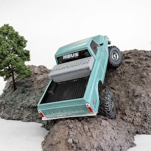 May include: A teal-colored toy pickup truck is angled on a small dirt mound. The truck has black tires and the text "HEUS" and "ArmorBuilt RC" on the vehicle. Two small model trees are also visible.