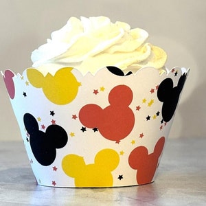 May include: A cupcake in a decorative paper liner featuring Mickey Mouse silhouettes in red, yellow, and black, with star accents. The cupcake has a swirl of white frosting.