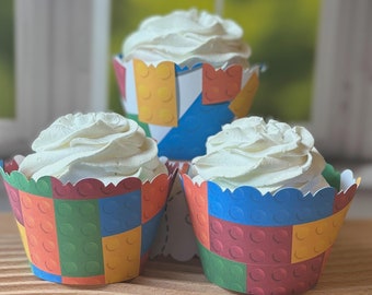 May include: Three cupcakes with white frosting sit in colorful paper wrappers. The wrappers are designed to resemble toy building blocks in red, yellow, blue, and green. The cupcakes are arranged on a wooden surface.