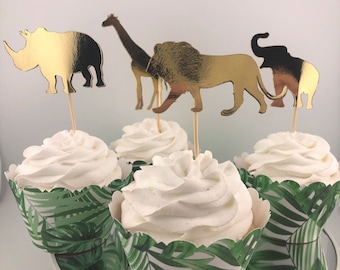 Pode incluir: Cupcakes com cobertura branca e enfeites dourados de animais, incluindo um rinoceronte, uma girafa, um leão e um elefante. As forminhas de cupcake apresentam um padrão de folhas tropicais verdes.