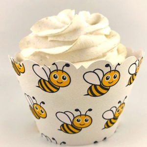 May include: A cupcake with white frosting sits in a decorative paper liner. The liner is white with a repeating pattern of cartoon bees in yellow and black. The cupcake is a dessert.