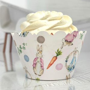 May include: A cupcake in a decorative paper cup with a scalloped edge. The cup features illustrations of Peter Rabbit, carrots, and floral elements. The cupcake is topped with a swirl of white frosting.