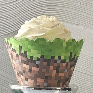 May include: A cupcake with white frosting sits in a decorative paper cup. The cup features a pixelated design with brown, green, and gray squares, resembling a video game theme. The cupcake is on a silver pedestal.