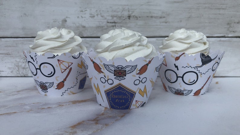 May include: Three cupcakes with white frosting in decorative paper cups. The cups feature a repeating pattern of glasses, a broom, an owl, and lightning bolts. One cup has the text "Chocolate Frog".