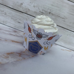 May include: A cupcake with white frosting sits in a decorative paper wrapper. The wrapper features illustrations of an owl, glasses, a broom, and lightning bolts. The wrapper also has the text "Chocolate Frog".