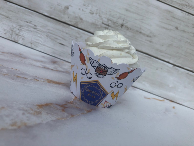 May include: A cupcake with white frosting sits in a decorative paper wrapper. The wrapper features illustrations of an owl, glasses, a broom, and lightning bolts. The wrapper also has the text "Chocolate Frog".