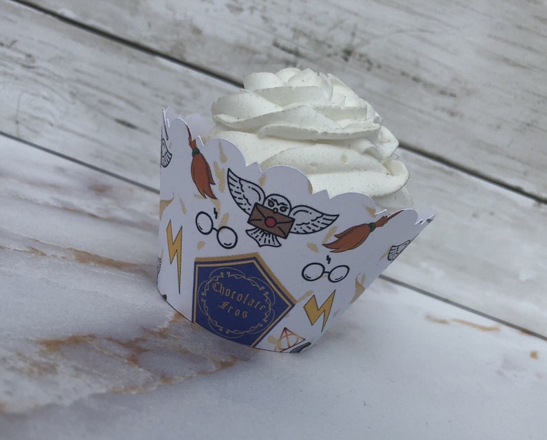 May include: A cupcake with white frosting sits in a decorative paper wrapper. The wrapper is white with repeating illustrations of a broom, owl, glasses, and lightning bolt. The wrapper also has the text "Chocolate Frog".