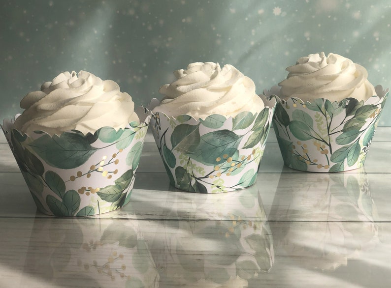 May include: Three cupcakes with white frosting in decorative paper cases. The cases have a green and gold leaf pattern. The cupcakes are arranged on a light-coloured wooden surface.