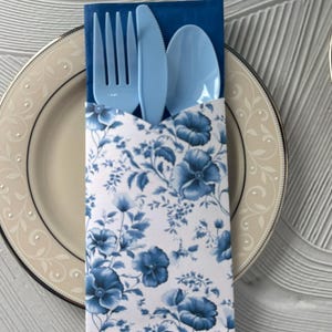 May include: A place setting with a blue plastic fork, knife, and spoon in a floral print paper sleeve. The sleeve features a blue and white floral pattern. The plate has a cream-colored rim with a decorative design.