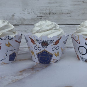 May include: Three cupcakes with white frosting in decorative paper cups. The cups feature a white background with repeating illustrations of glasses, lightning bolts, brooms, and the text "Chocolate Frog".