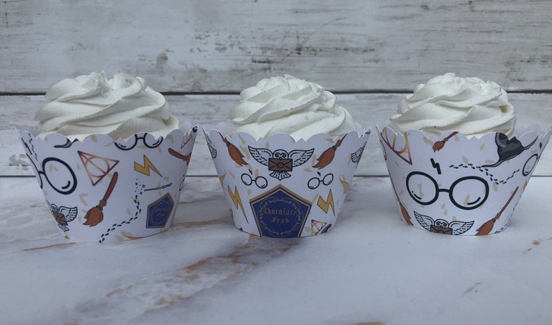 May include: Three cupcakes with white frosting in decorative paper cups. The cups feature a white background with repeating illustrations of glasses, lightning bolts, brooms, and the text "Chocolate Frog".